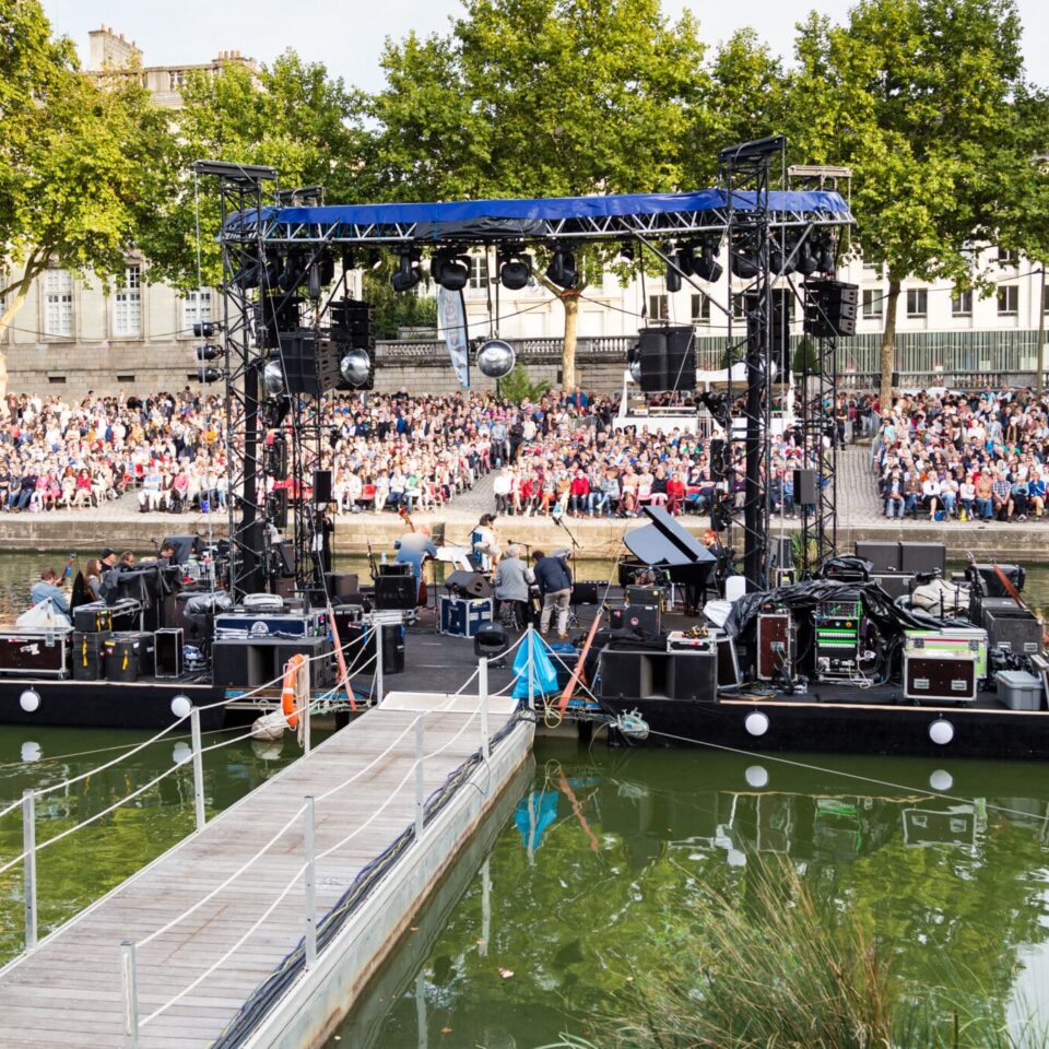 Spectacle musical sur une scène flottant sur une rivière, avec nombreux spectateurs sur des gradins. RDV de l'Erdre, Nantes